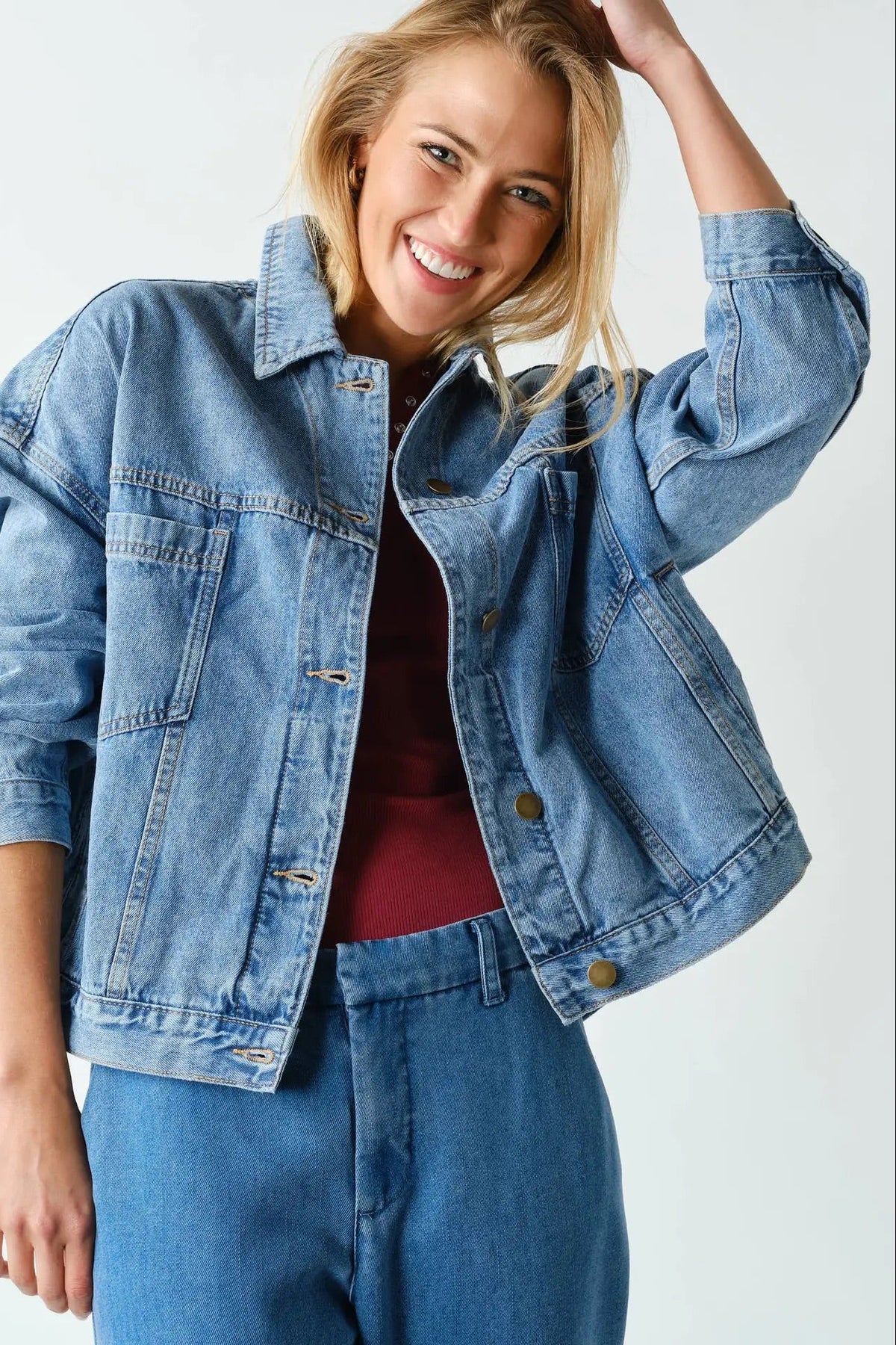 A smiling blonde woman wearing a cropped light-wash blue denim jacket over a burgundy top and blue jeans, posed against a clean white studio background.