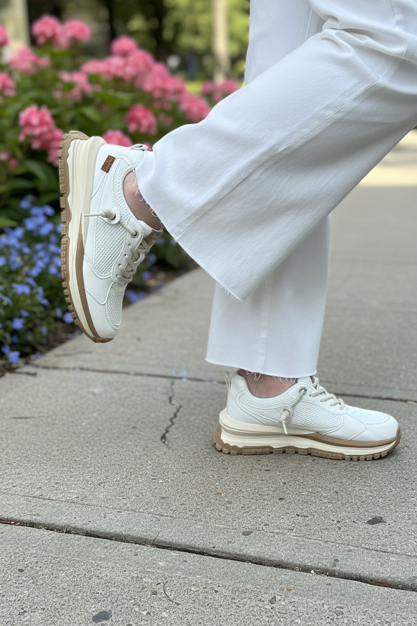 White sneakers with brown soles worn by a person standing on a sidewalk with flowers in the background.
