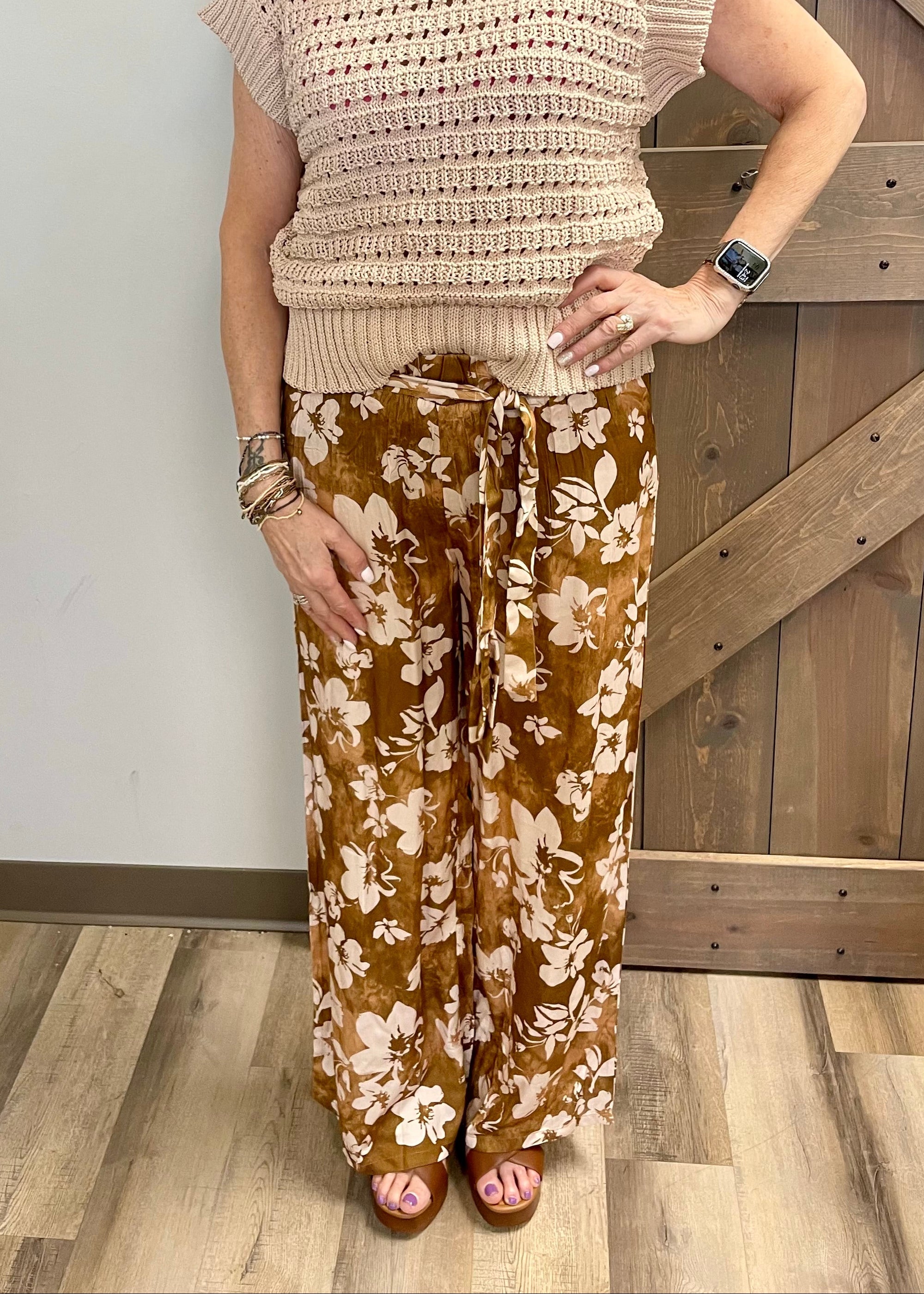 Woman wearing a beige crochet top and floral pants standing in front of a wooden door.
