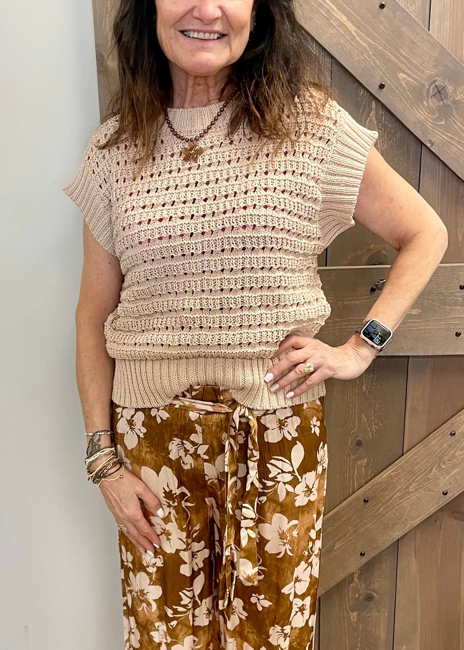 Woman wearing a beige crochet top and floral pants standing in front of a wooden door.