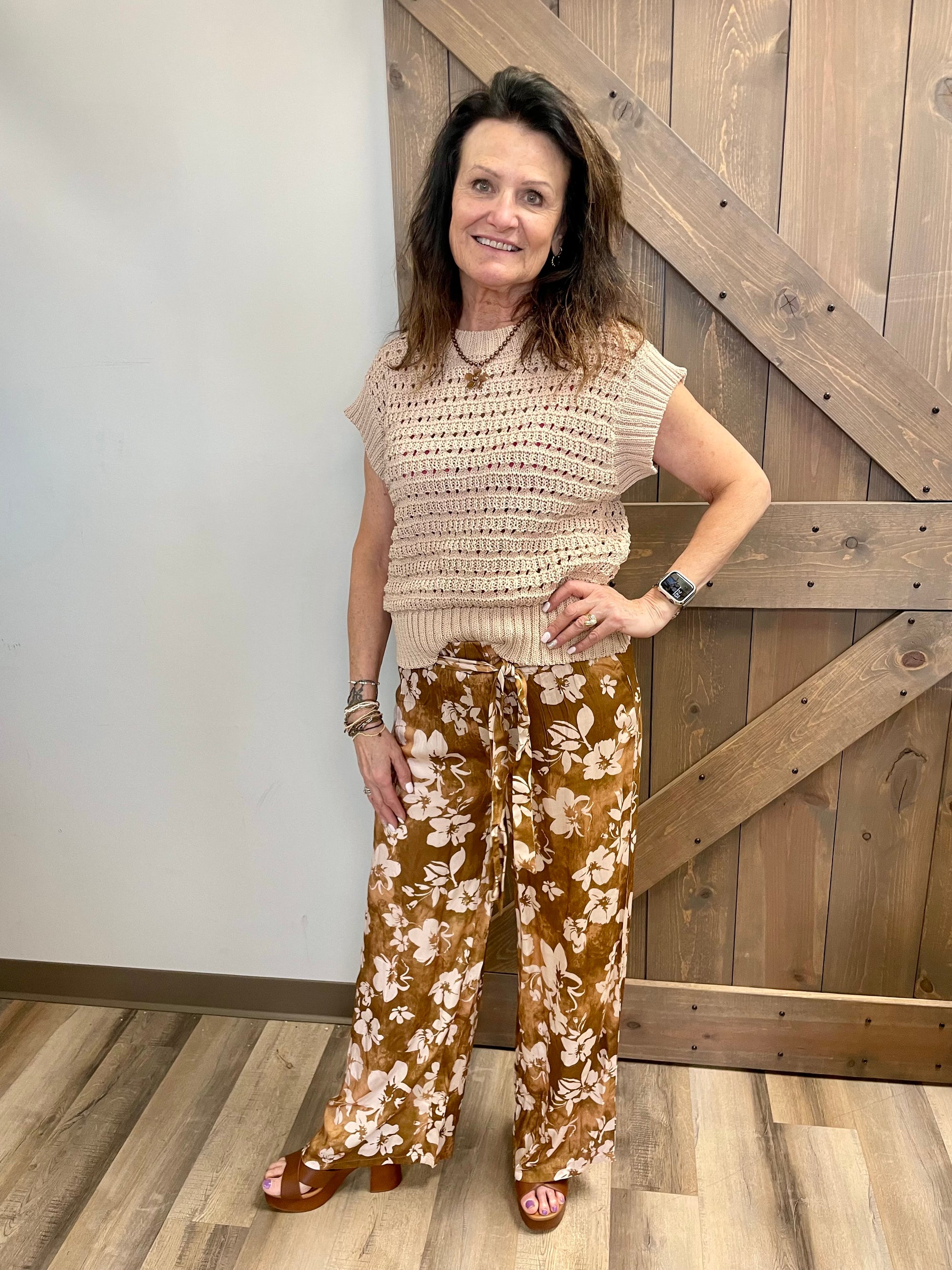 Woman wearing a beige sleeveless top and brown floral pants standing in front of a wooden door.