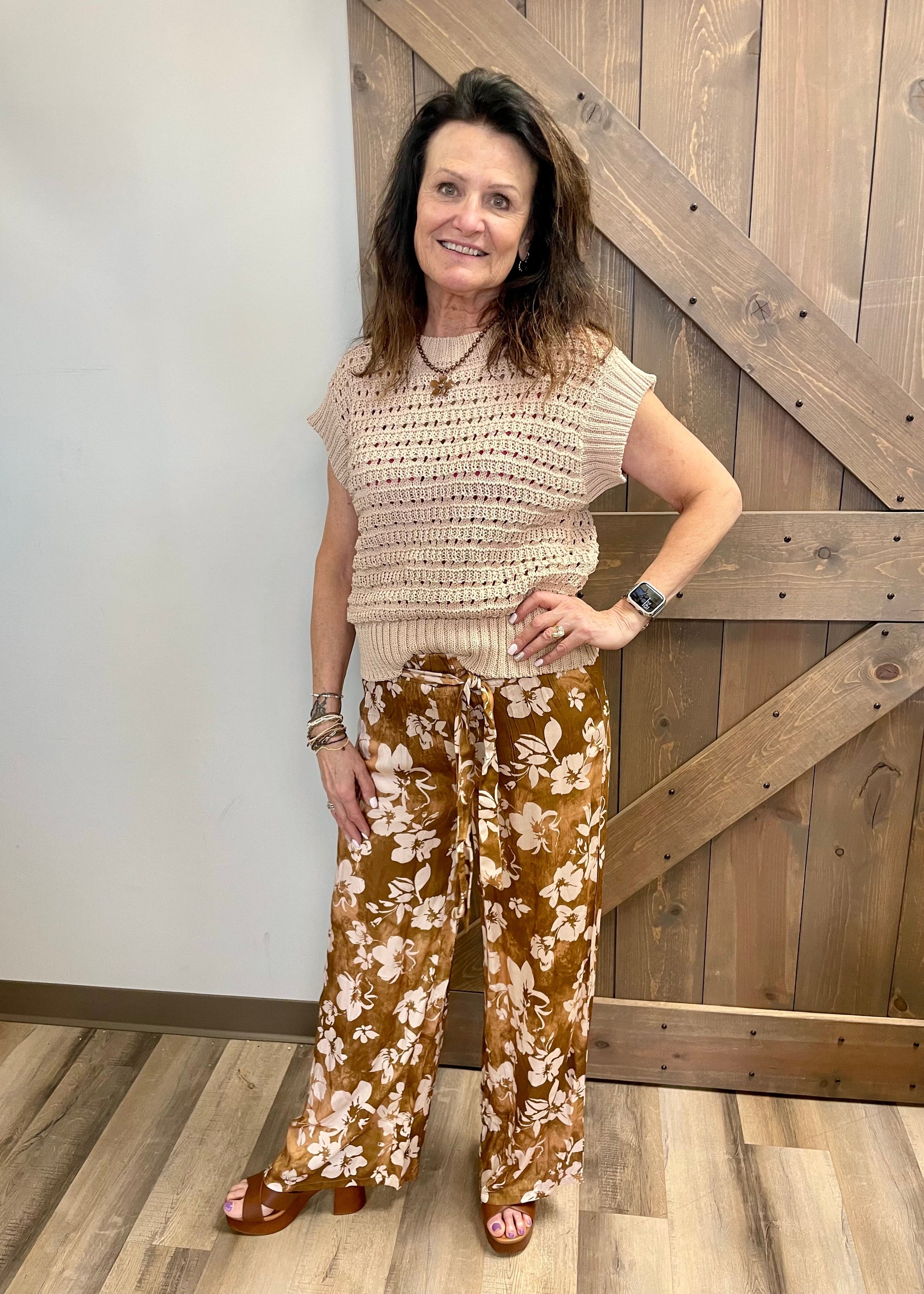Woman wearing a beige sleeveless top and brown floral pants standing in front of a wooden door.