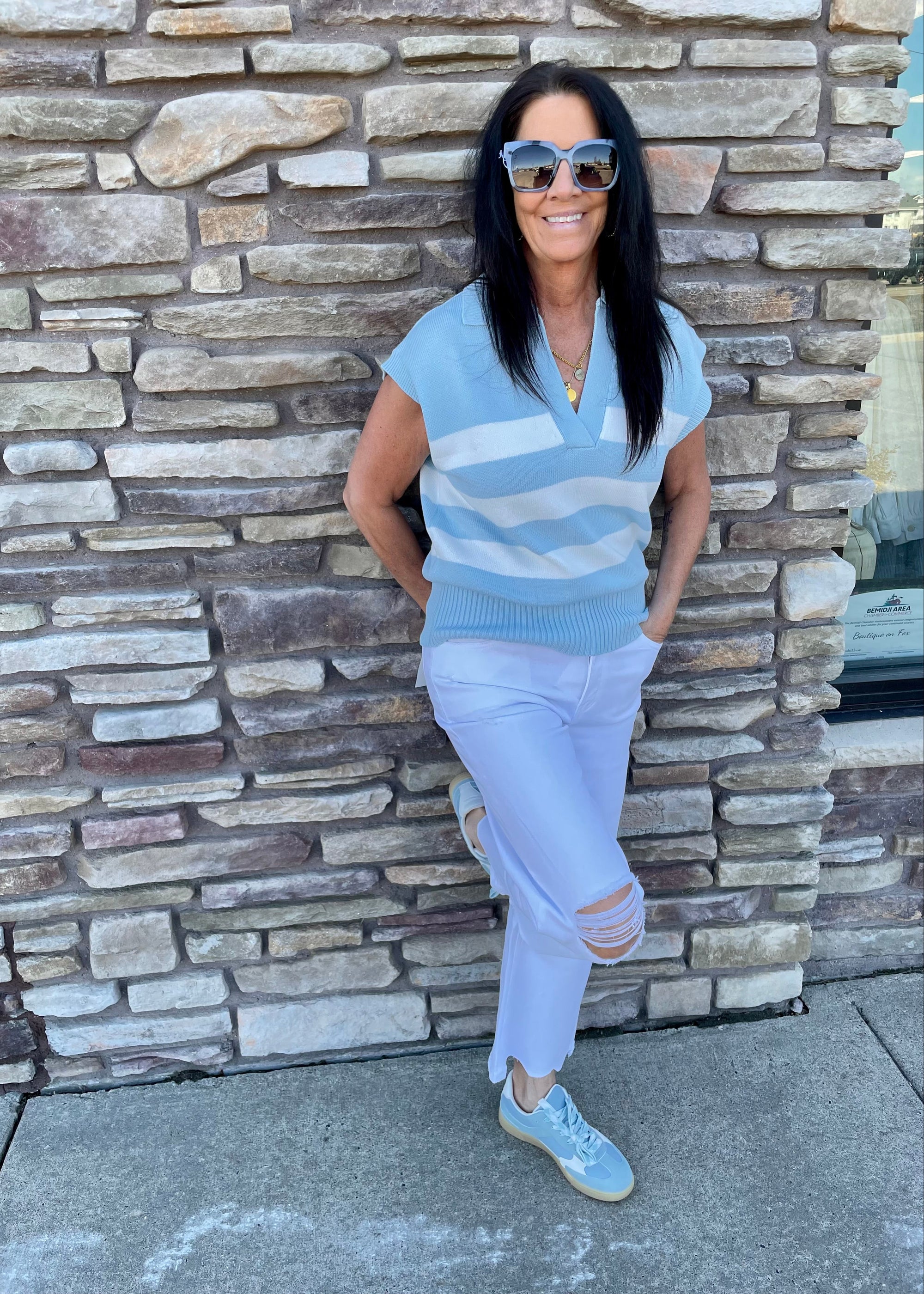 Woman wearing a light blue striped sweater and white pants leaning against a stone wall.