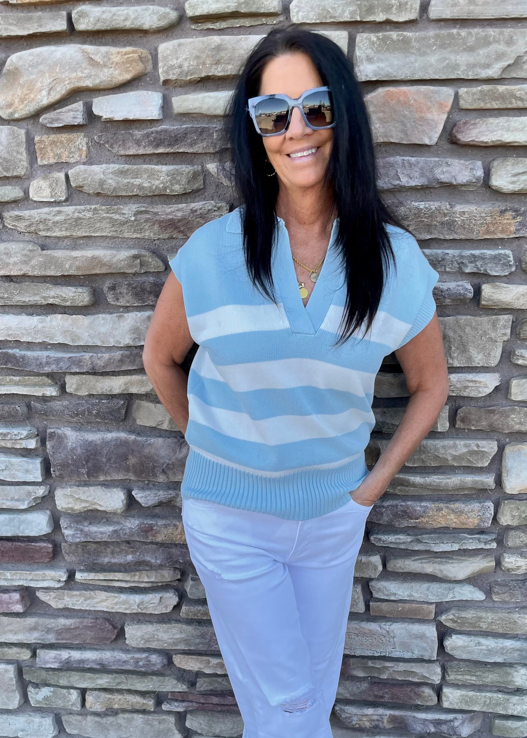 Woman wearing a blue striped shirt and white jeans standing against a stone wall.
