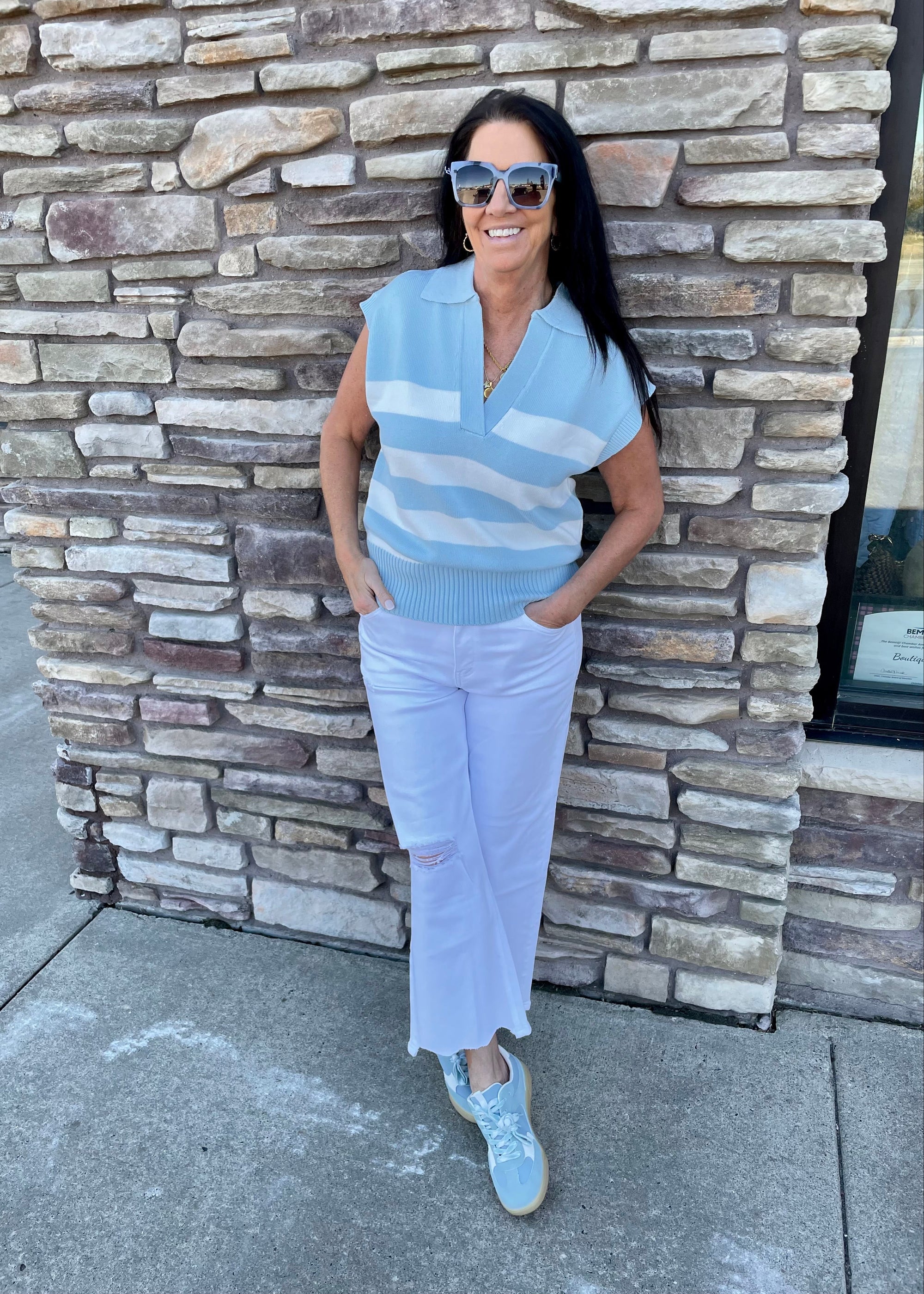 Woman wearing a light blue striped sleeveless top and white pants standing against a stone wall.