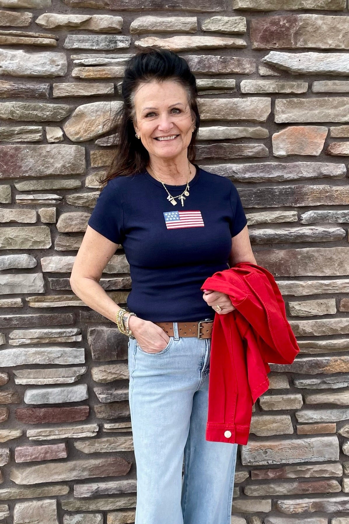 Woman wearing a blue t-shirt with an American flag design, light blue jeans, and red shoes, standing against a stone wall.