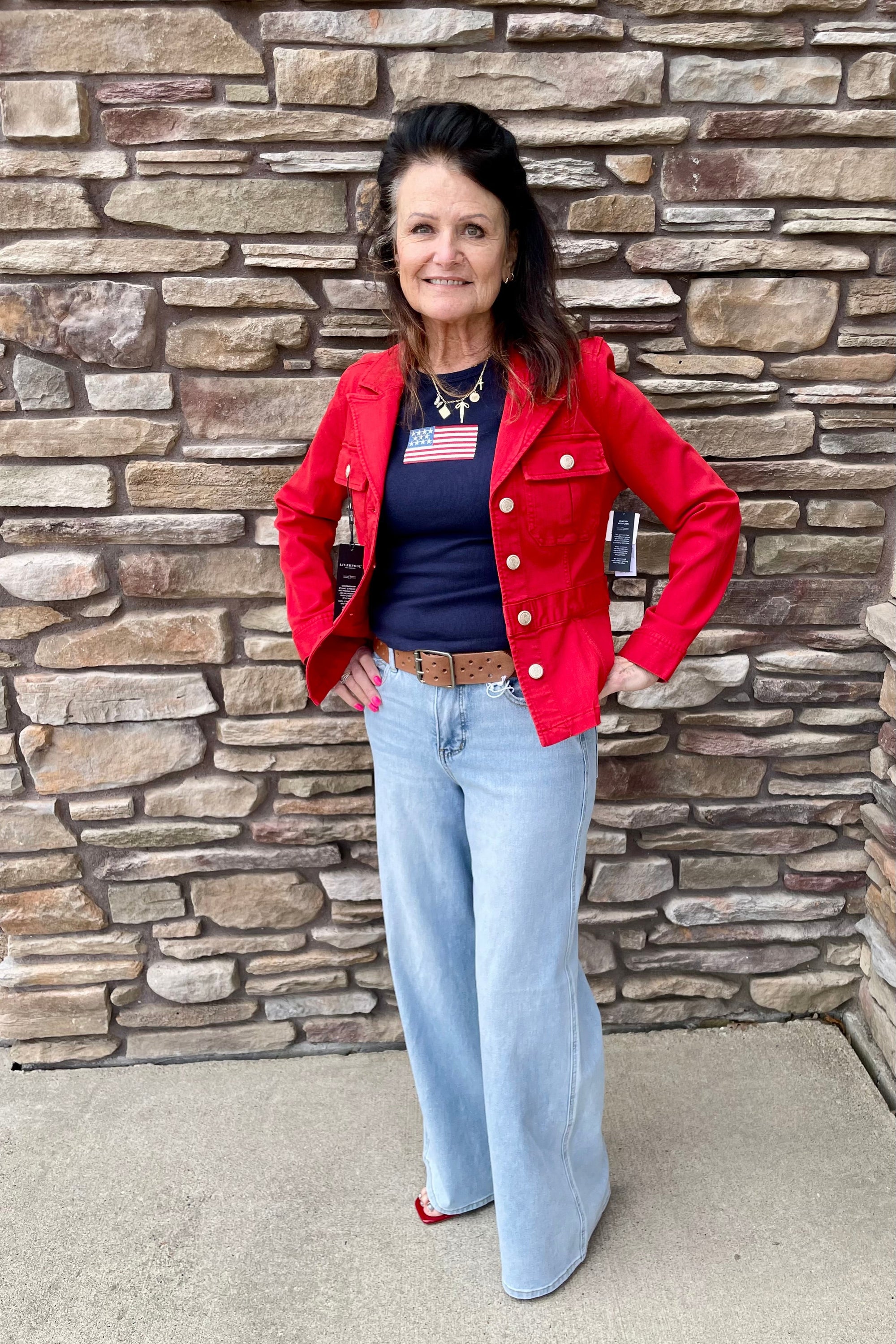 Woman wearing a red jacket, blue jeans, and an American flag shirt standing in front of a stone wall.