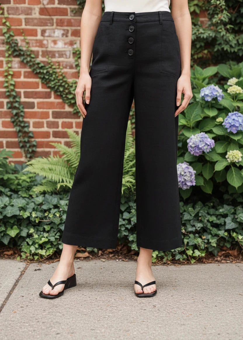 Person wearing black wide-leg pants standing in front of a brick wall and greenery.