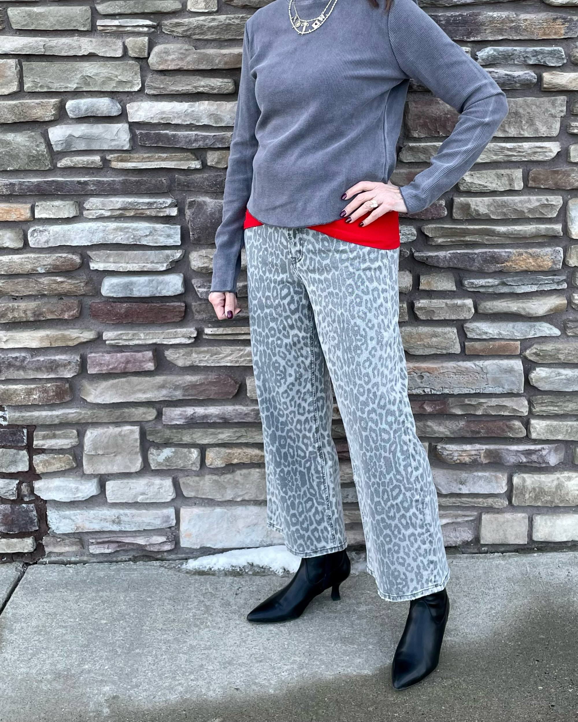 Person wearing a gray sweater, red shirt, white pants with black leopard print, and black boots against a stone wall.