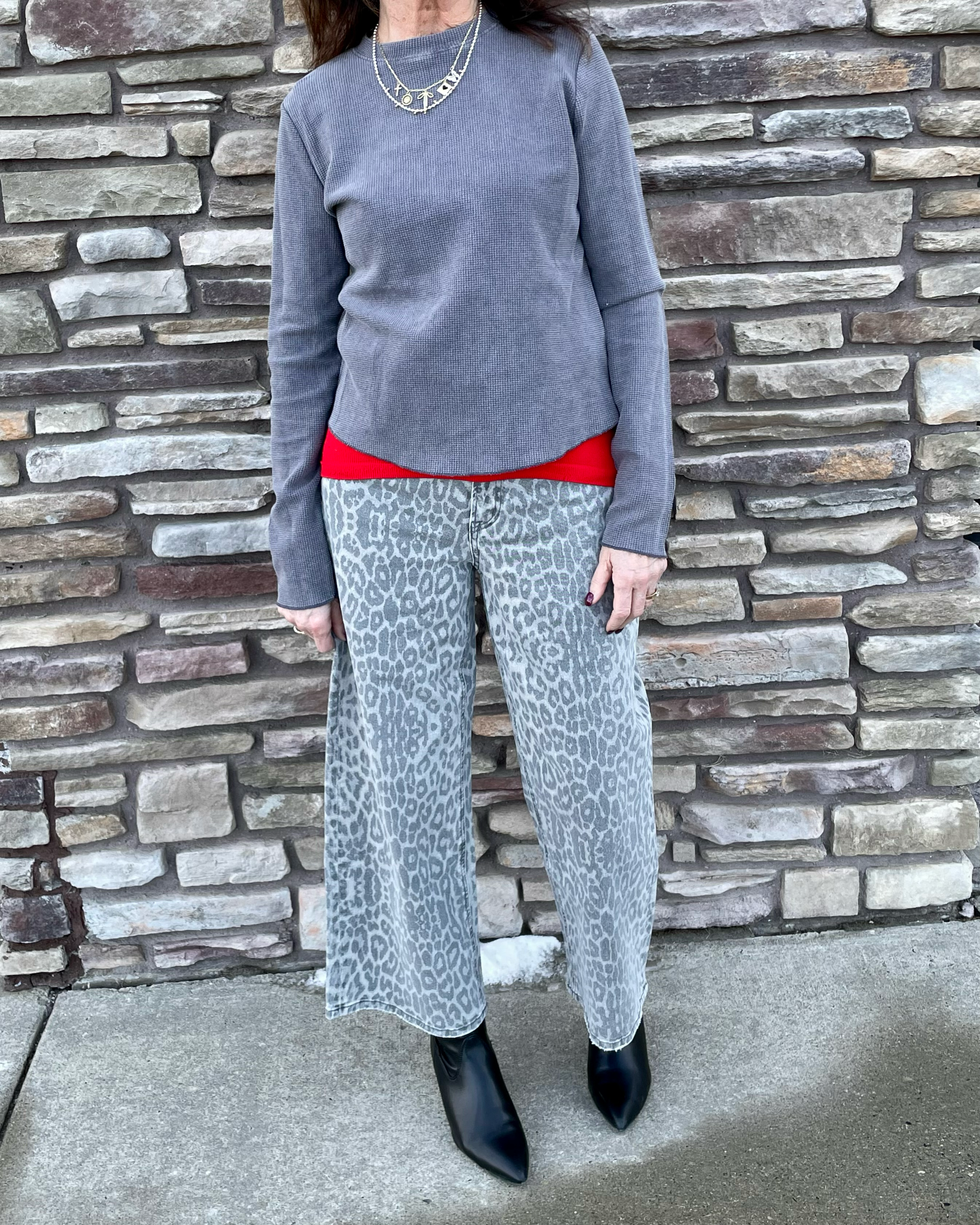 Person wearing a gray sweater, red shirt, and leopard print pants against a stone wall.