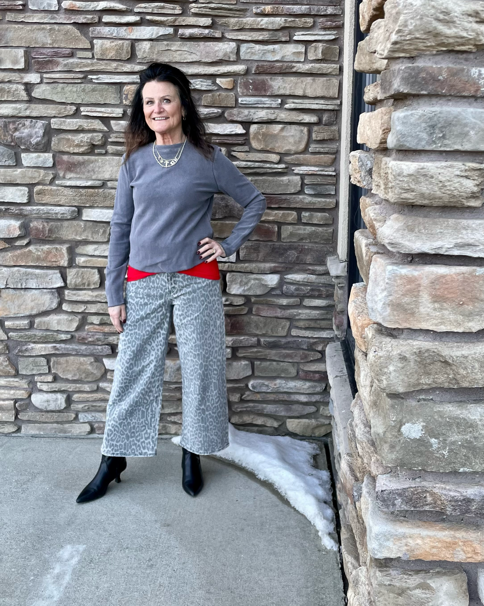 Woman in gray sweater and patterned pants standing against a stone wall.